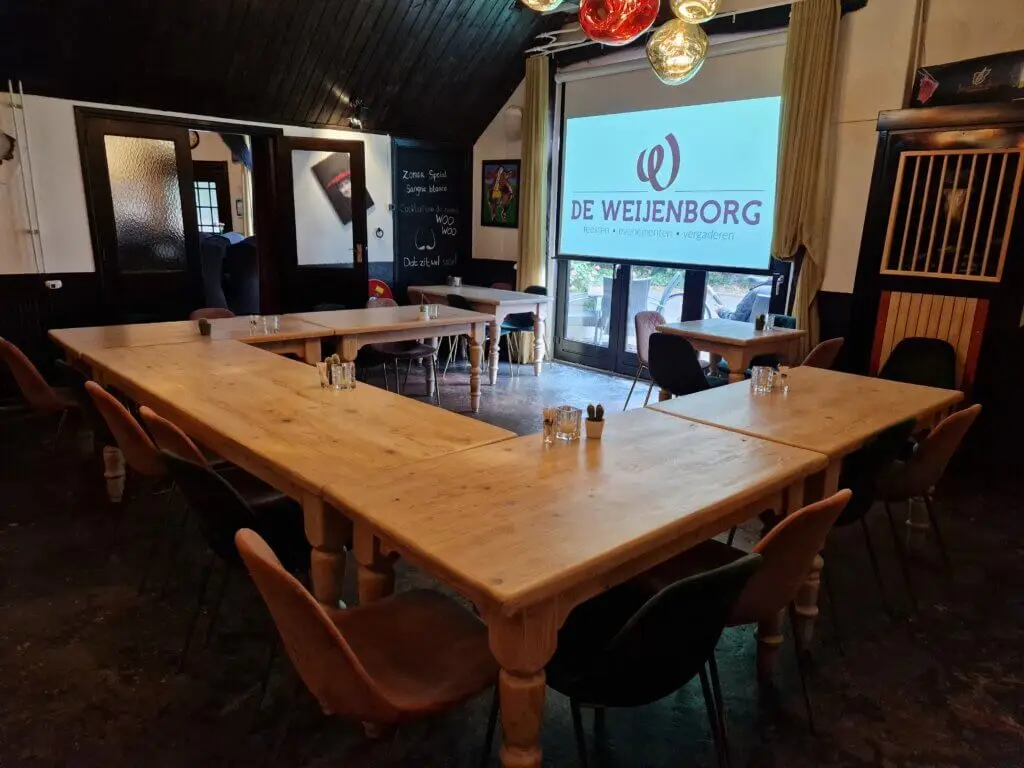 Vergaderzaal met houten tafels in U-opstelling en een projectiescherm bij De Weijenborg in Delden